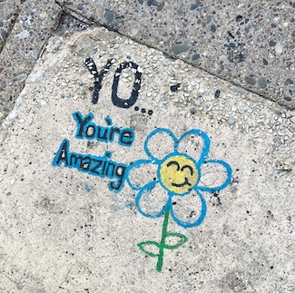 a positive message of "you're amazing" with a smiling flower drawn on a city sidewalk