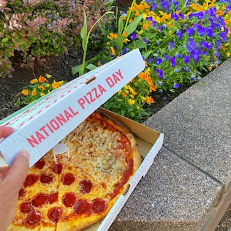 a fresh pizza with pepperoni in a box outside next to pretty colorful flowers