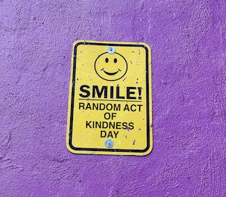 a bright yellow metal sign on a purple wall asking you to smile for random act of kindness day in February