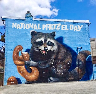 a blue mural on an old building of a raccoon holding a pretzel on a bright sunny day