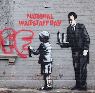 a famous street art (graffiti) artist rendition of a young man spray painting a brick wall in a city, with a waiter holding a try with either two paint cans...or two martini shakers...for national waitstaff day in May.
