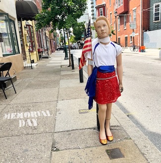 a female mannequin dressed in a red skirt, white top and a blue sash, holding an American UISA flag on a. city street for Memorial Day