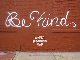 white graffiti of "BE KIND" spray painted on a red brick wall for world kindness day