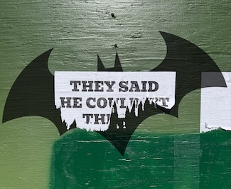 a torn paper poster on a green plywood wall, with the batman symbol