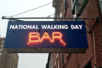 a neon bar sign lit, hanging from a pole in a city, for national walking day