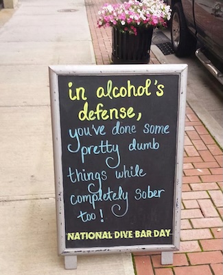 a fun, cursive written street sign about drinking, outside a bar on a city street
