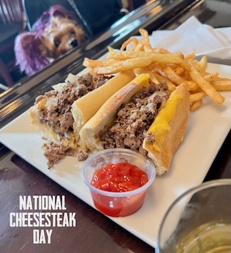 A tasty cheesesteak with fries on a bar with a small puppy looking at it