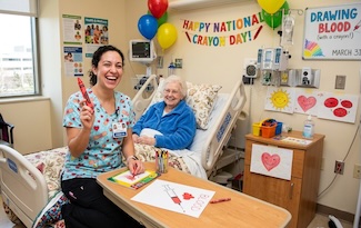 a nurse and elderly patient in a hospital bed. The nurse is holding a big red crayon to "draw blood" for national crayon day.