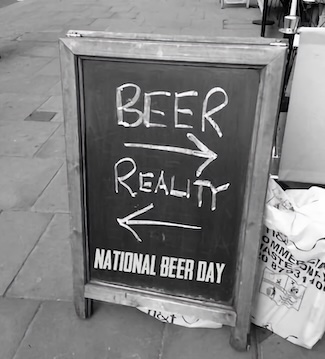 A black and white photo of a sidewalk A-frame chalkboard sign. The hand-written text reads: 'BEER' with an arrow pointing right, and 'REALITY' with an arrow pointing left. Bold text at the bottom reads: 'NATIONAL BEER DAY'.
